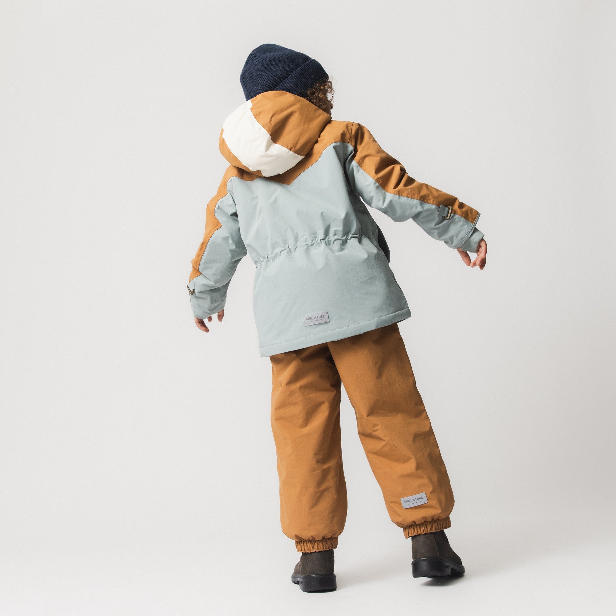 Et barn set skråt bagfra iført en MATWALDO fleeceforet vinterjakke med hætten oppe, stylet med matchende gyldenbrune vinterbukser og en mørkeblå hue. Jakkens colorblock-design i støvet mintgrøn og gyldenbrun med hvid hætte er tydeligt synlig, mens barnet strækker armen ud til siden.