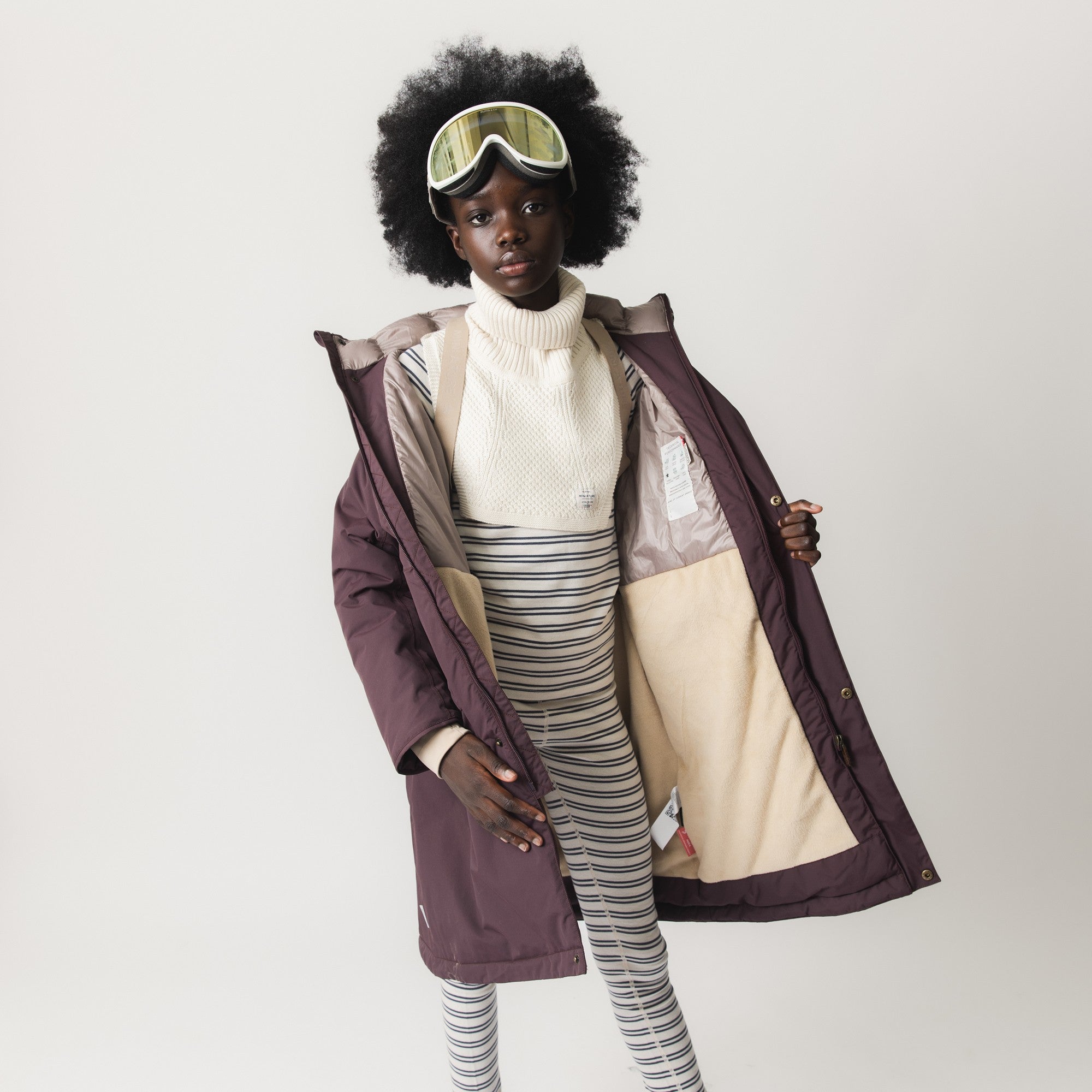 Pigen har MATVENCASIA-fleeceforet vinterjakke i mørk bordeaux åben og viser det beige fleecefór og de indvendige seler. Hun er stylet med en lys strikket halsvarmer, stribede leggings og skibriller.