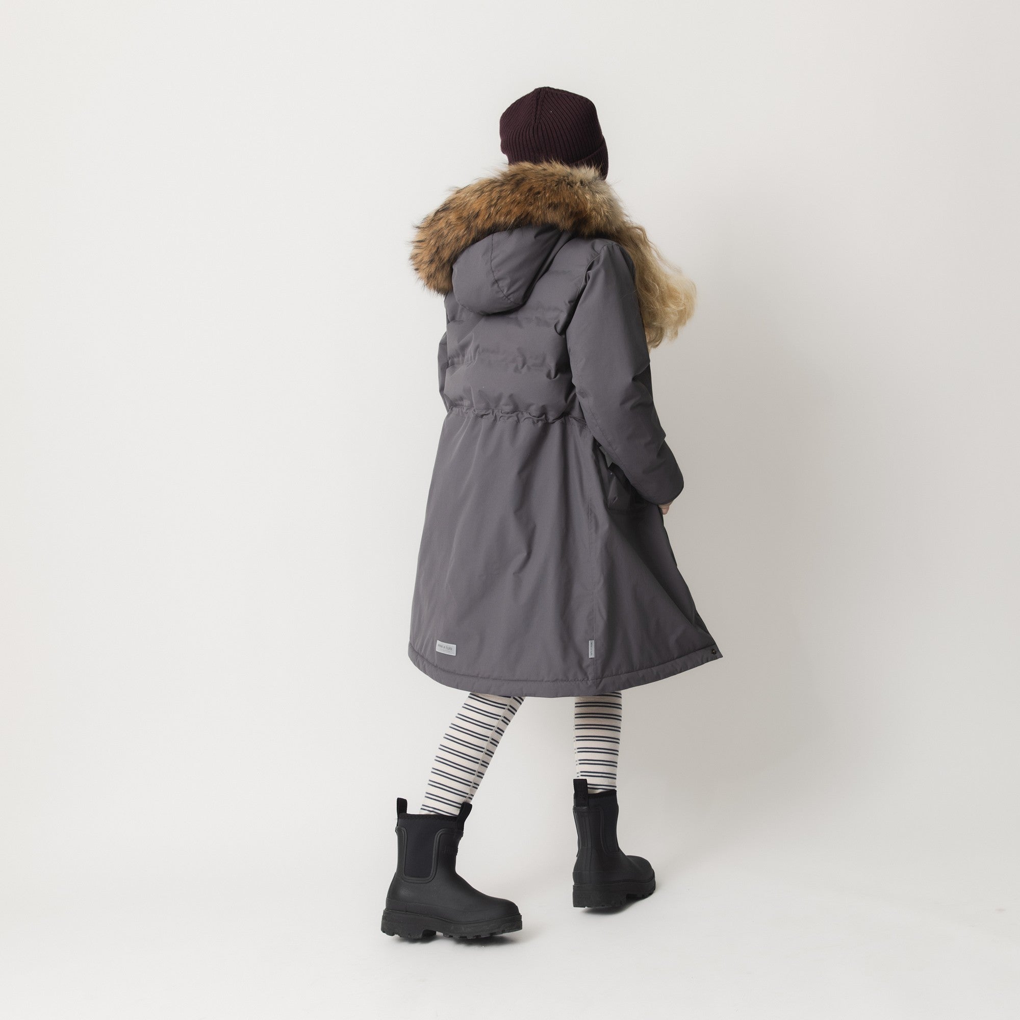 Set bagfra fremhæver MATVENCASIA’s fleeceforede vinterjakke med pels det lange snit ned over hofterne og den store, aftagelige pelskant på hætten i en naturlig brun nuance. Jakken er stylinget med en mørk hue, stribede leggings og sorte gummistøvler mod en lys baggrund.