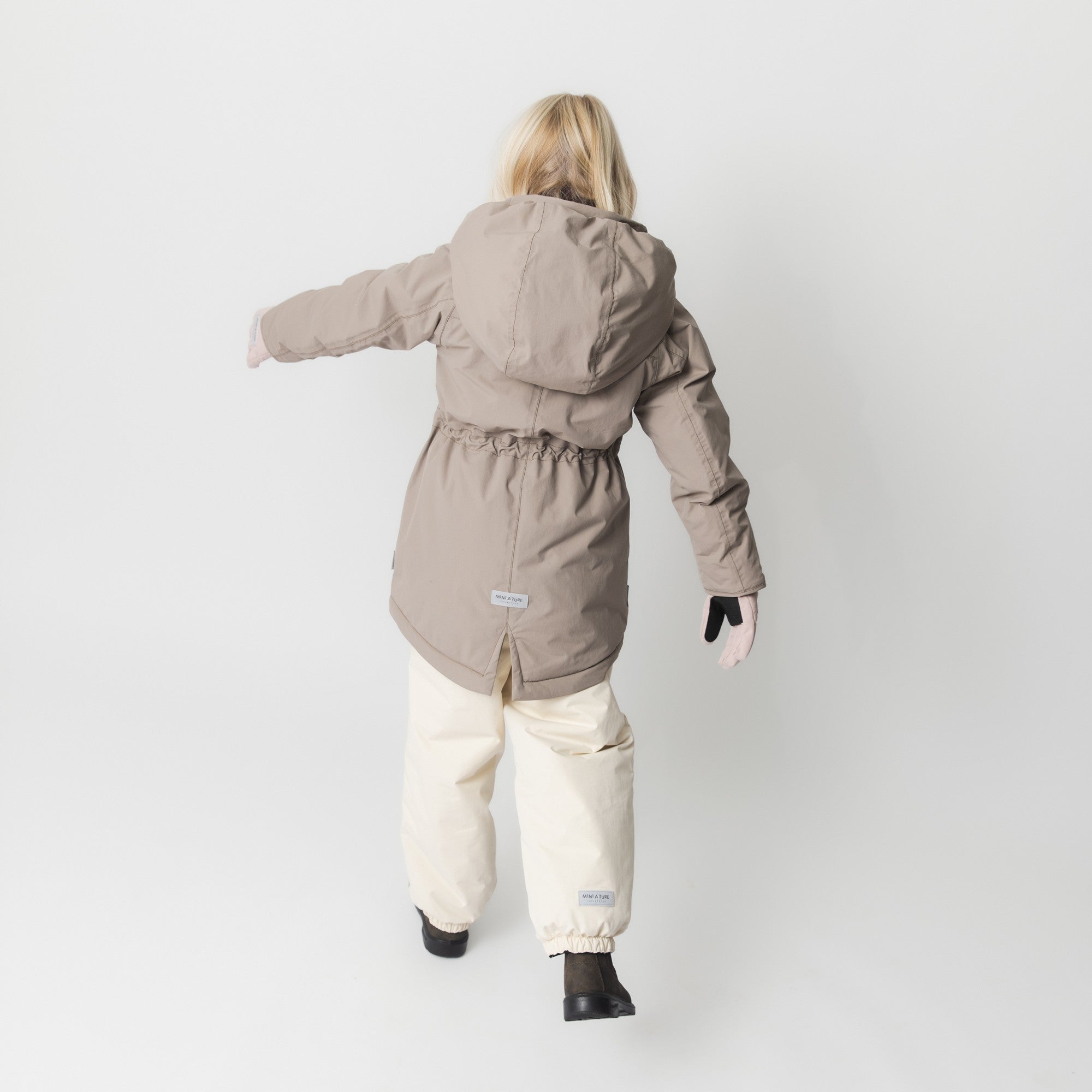 Set bagfra viser modellen MATVIKANIA-fleeceforet vinterjakke i sandbeige med tydelig elastik i taljen og den karakteristiske, længere model. Jakken er kombineret med cremehvide vinterbukser og mørke støvler.