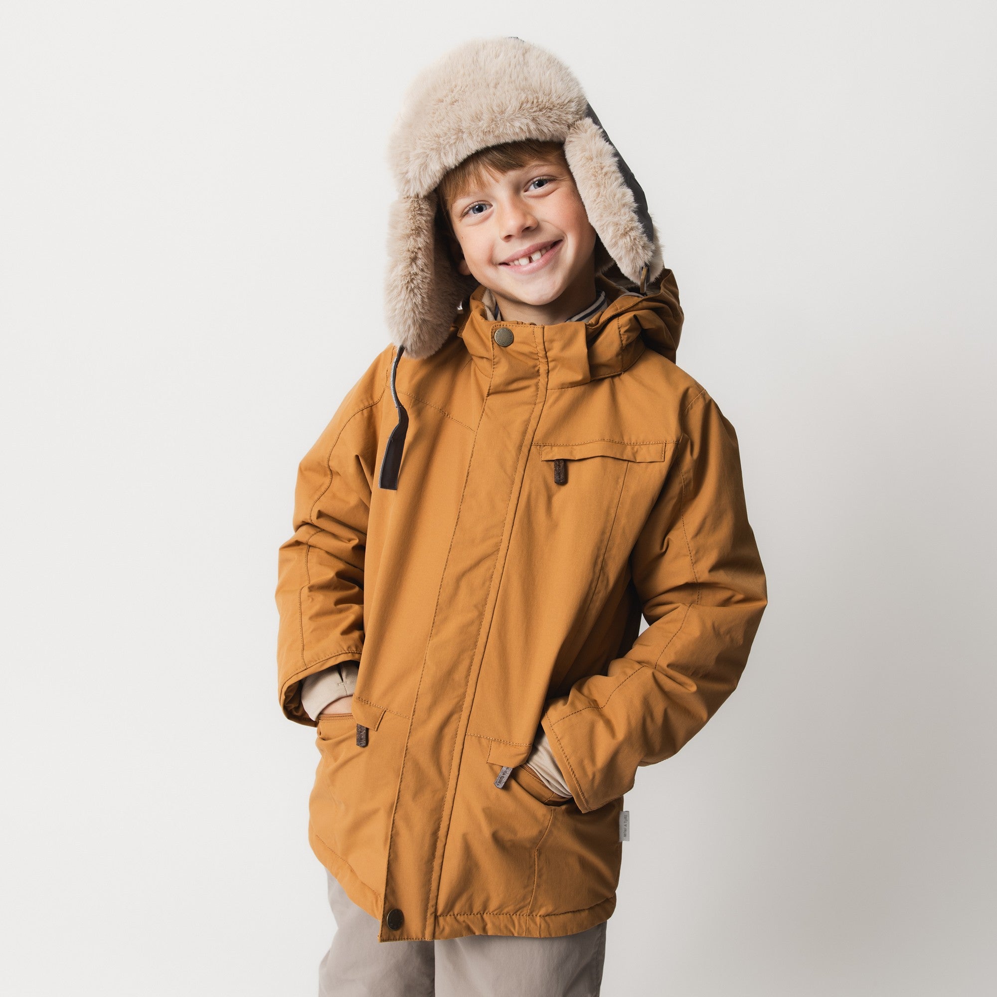 Et smilende barn iført MATVESTYN vinterjakke i en varm okkerfarvet nuance med hænderne i de nedre frontlommer. Jakken er kombineret med en beige pelsforet hue med trappeudskæringer og lyse bukser på en lys baggrund. Brystlommen med lynlås og den skjulte frontlynlås med trykknap øverst er tydeligt synlige.