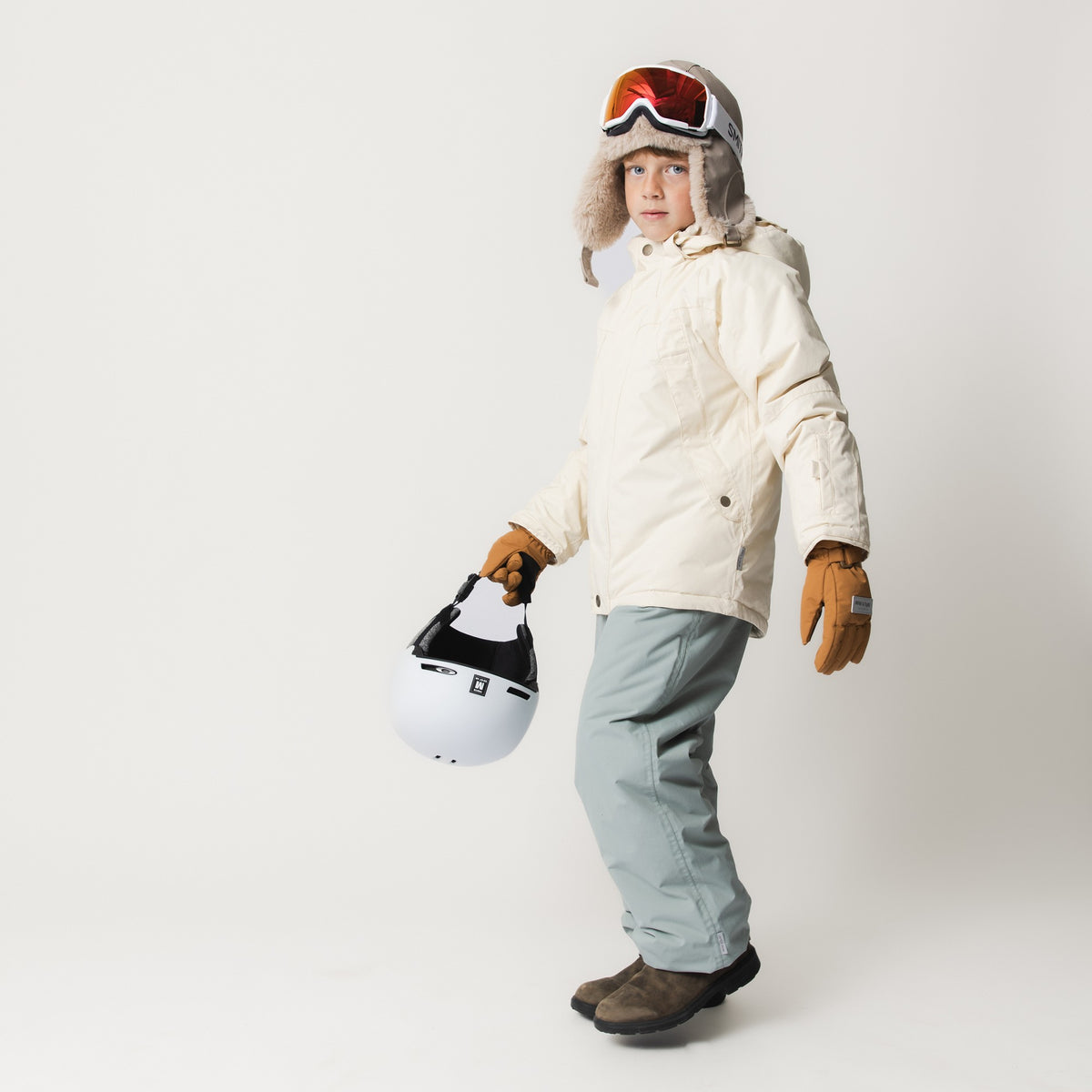 Et barn fotograferet i fuld figur fra siden iført en MATVALONSO skijakke i creme med hætten nede, kombineret med lysegrå skibukser, brune skihandsker og mørke støvler. Barnet har røde skibriller på en pelshat og holder en hvid skihjelm i hånden.