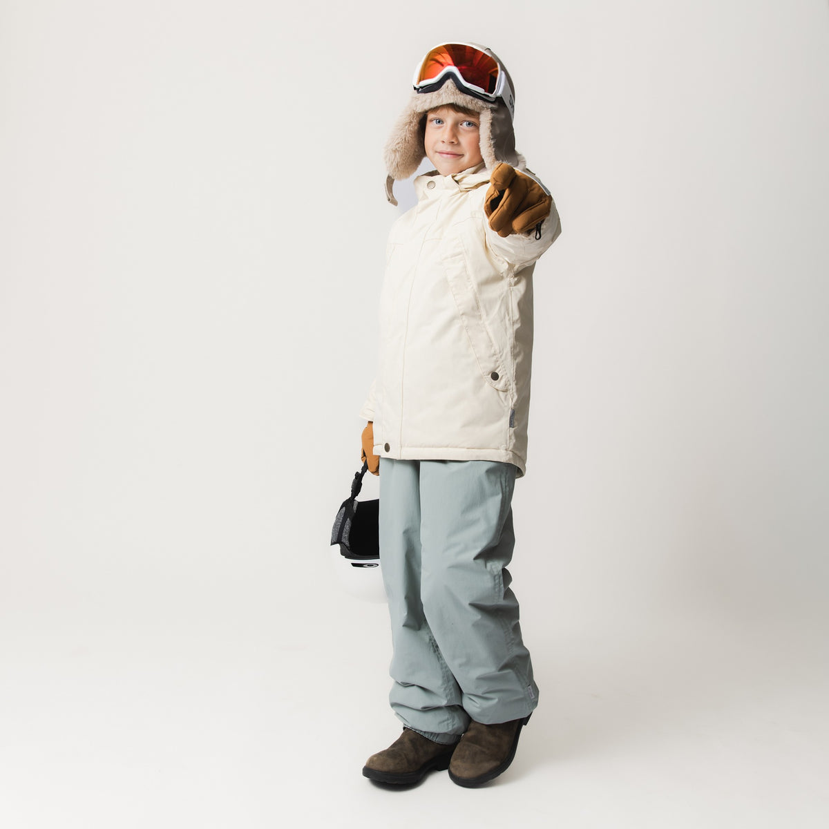 Barnet er fotograferet i fuld figur iført en MATVALONSO-skijakke i ensfarvet creme, som er kombineret med lysegrå skibukser, brune skistøvler og en pelshat med skibriller. Barnet peger mod kameraet med en brun skihandske og holder et par ekstra skibriller i den anden hånd.