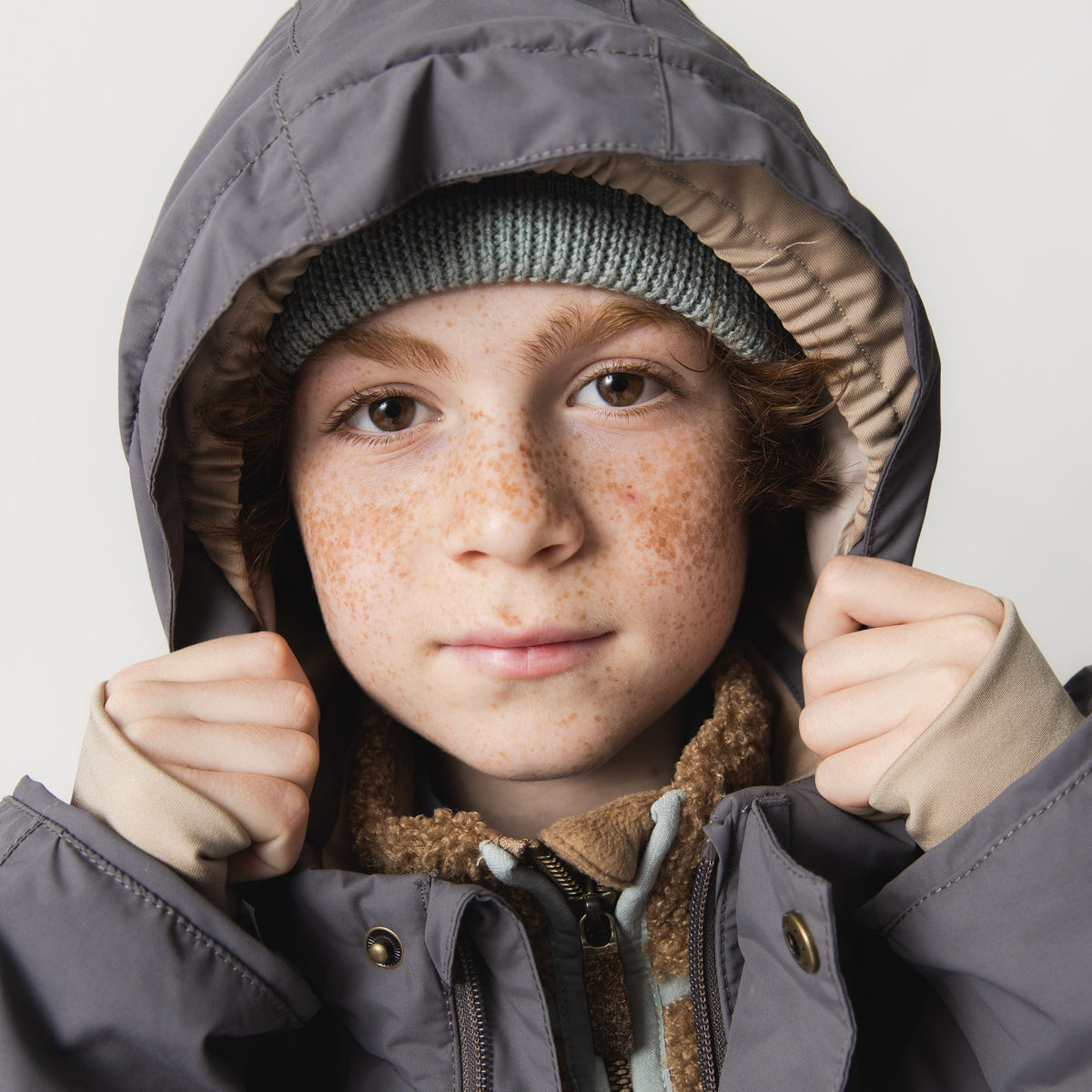 Nærbillede af et barn med rødt hår og fregner, der trækker hætten på en MATVALONSO-skijakke i gråt tæt om ansigtet med begge hænder. Under jakken ses en brun teddyfleece og en lysegrøn trøje, mens en grøn strikhue stikker frem inde i den cremefarvede hætte.