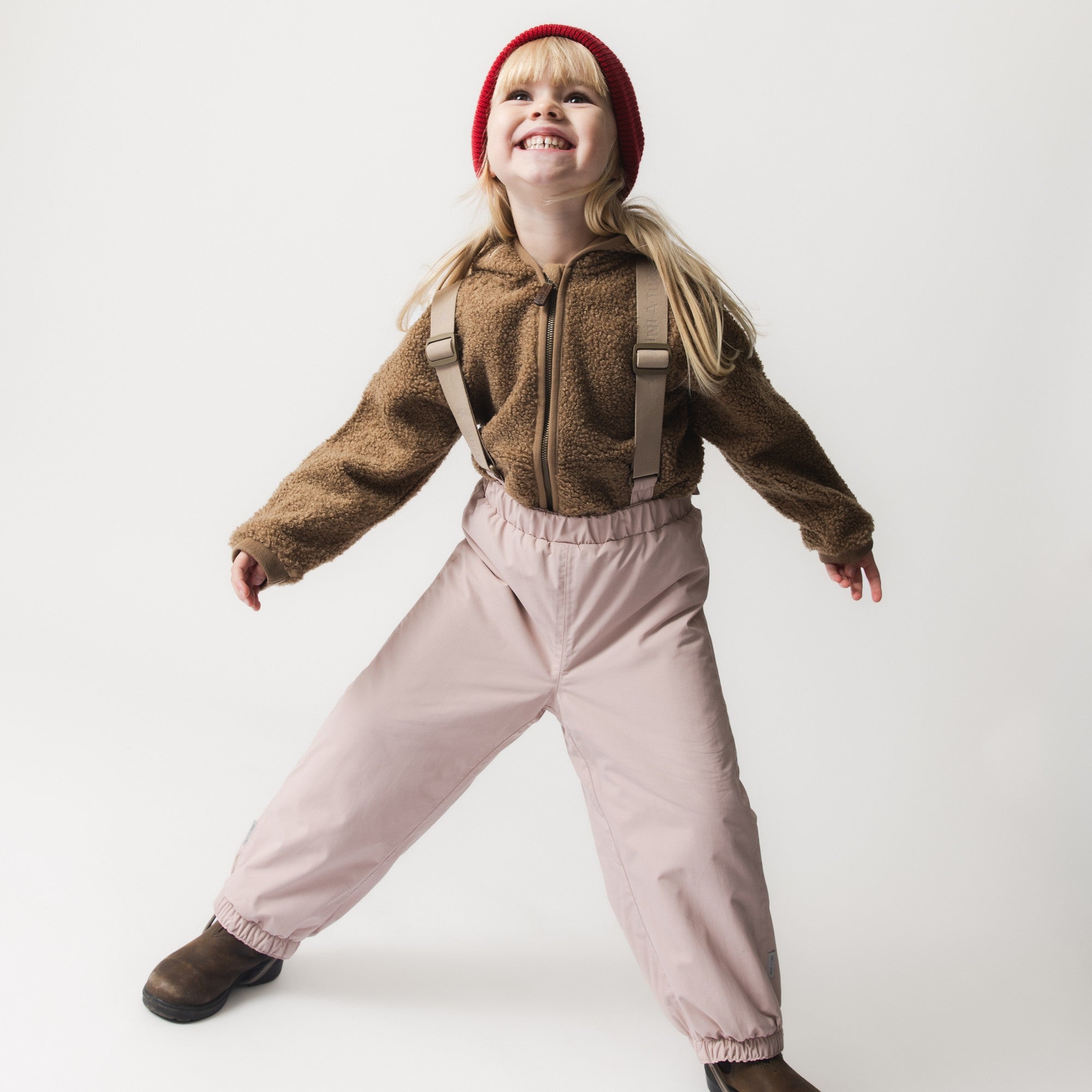 Et smilende barn iført MATWILAS overtræksbukser i pudderrosa med beige seler over en brun teddyfleece-jakke og rød hue, fotograferet i en aktiv positur mod hvid baggrund.