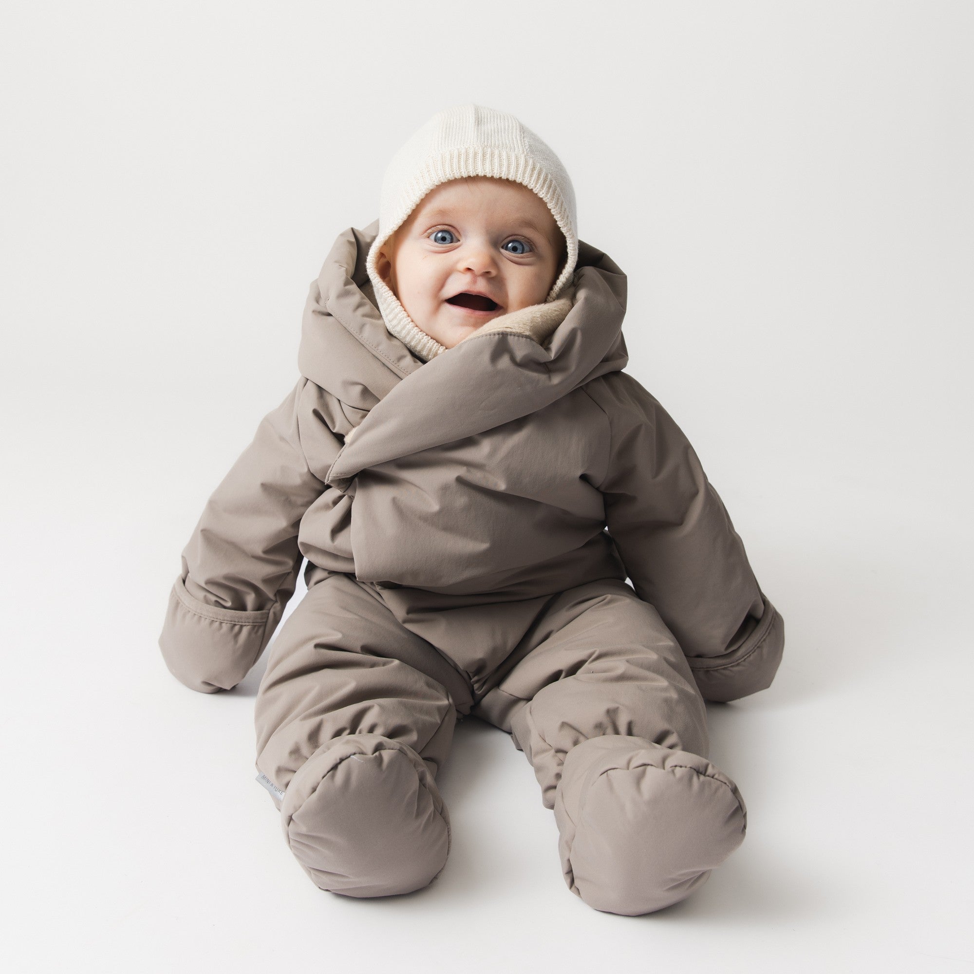 En glad baby sidder oprejst iført en MATFIANNA fleeceforet køredragt i varm gråbrun farve. Den diagonale trykknaplukning er delvist åben, så den skulderformede hætte folder sig ud omkring ansigtet. De integrerede luffer og futter er foldet op om hænder og fødder, og barnet har en cremefarvet strikket hue på.