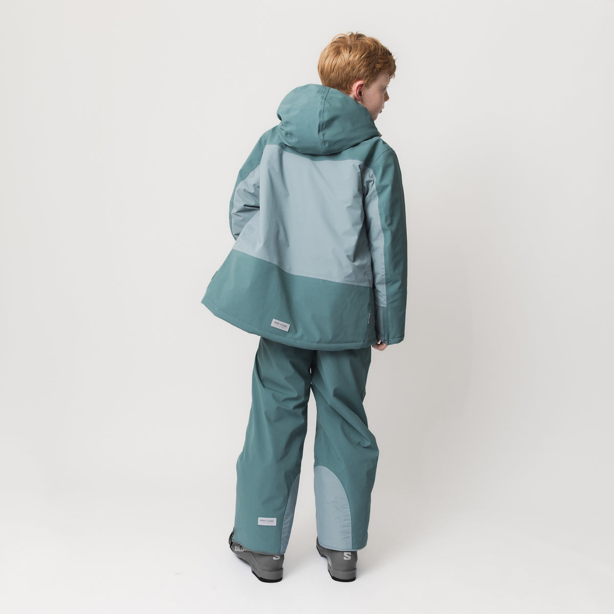 Barnet ses bagfra iført en MATADANE Ski Tech-jakke i blågrønt farveblokdesign med et lysegråt panel på ryggen, kombineret med matchende skibukser og skistøvler.
