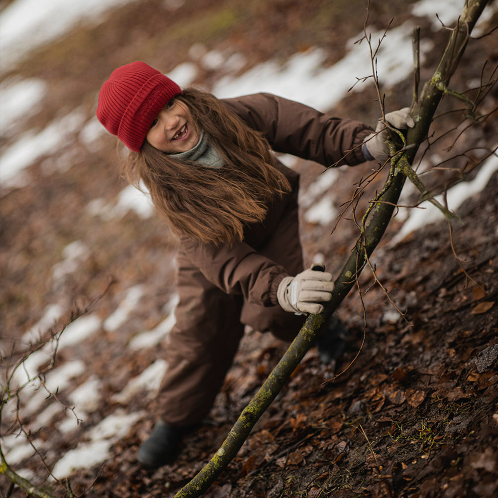 Et barn klatrer smilende op ad en skovskråning med sne i baggrunden iført en MATWANNI fleeceforet flyverdragt i chokoladebrun farve, rød strikhue og lysegrå vanter. Barnet holder fast i en gren for at komme op ad bakken.