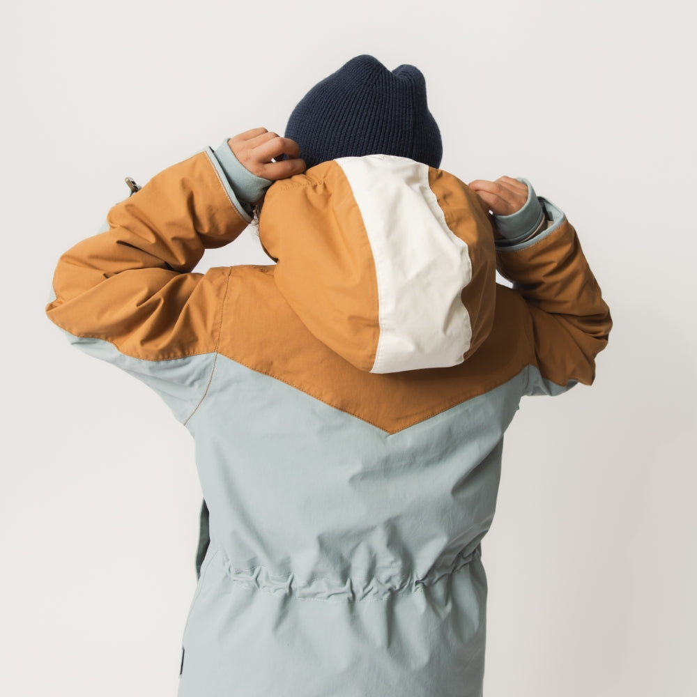 Et barn set bagfra iført en MATWALDO fleeceforet vinterjakke, mens det tager hætten af med begge hænder. Jakkens colorblock-design i støvet mintgrøn og gyldenbrun med hvid hætte samt de ribbede manchetter ved ærmerne er tydeligt synlige.