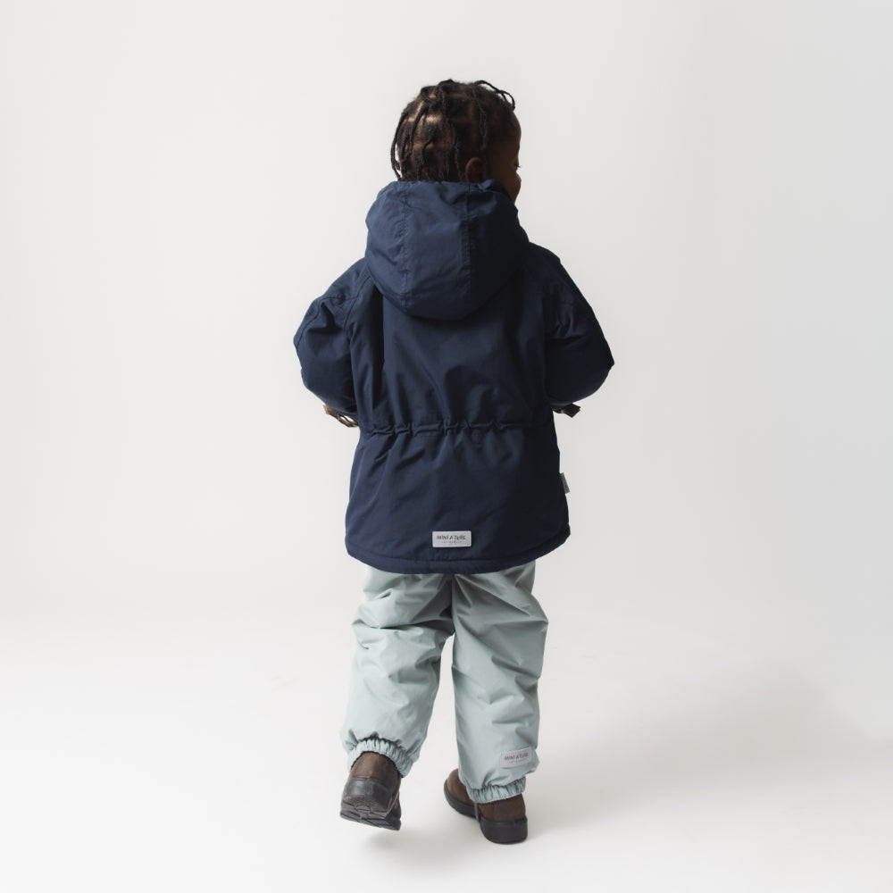 Et barn fotograferet bagfra i bevægelse iført en MATWALLY fleeceforet vinterjakke i mørk marineblå med hætten oppe. Den justerbare talje er synlig ved hoften. Outfittet er kombineret med lyse mintgrønne vinterbukser og mørkebrune støvler, og MINI A TURE-mærket ses centralt placeret nederst på ryggen.